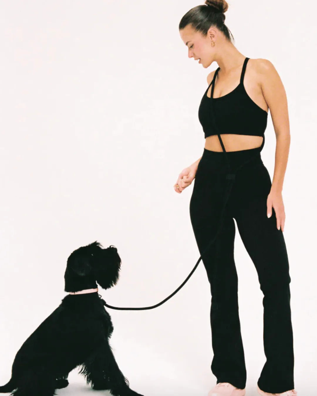 Woman in black athletic wear standing next to a small black dog on a white background with a luxury black rope dog lead