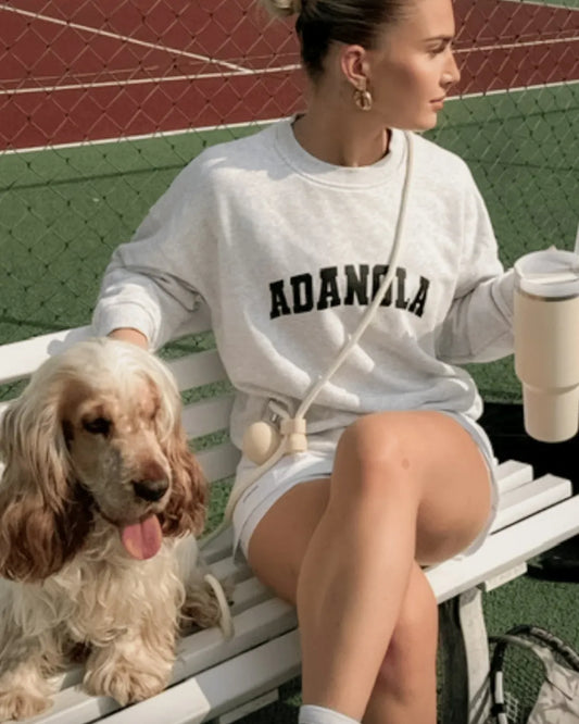 Woman sitting on a bench with a dog, wearing an 'ADANGELA' sweatshirt with a luxury handsfree lead around her body. She holds a neutral Stanley cup in one hand.
