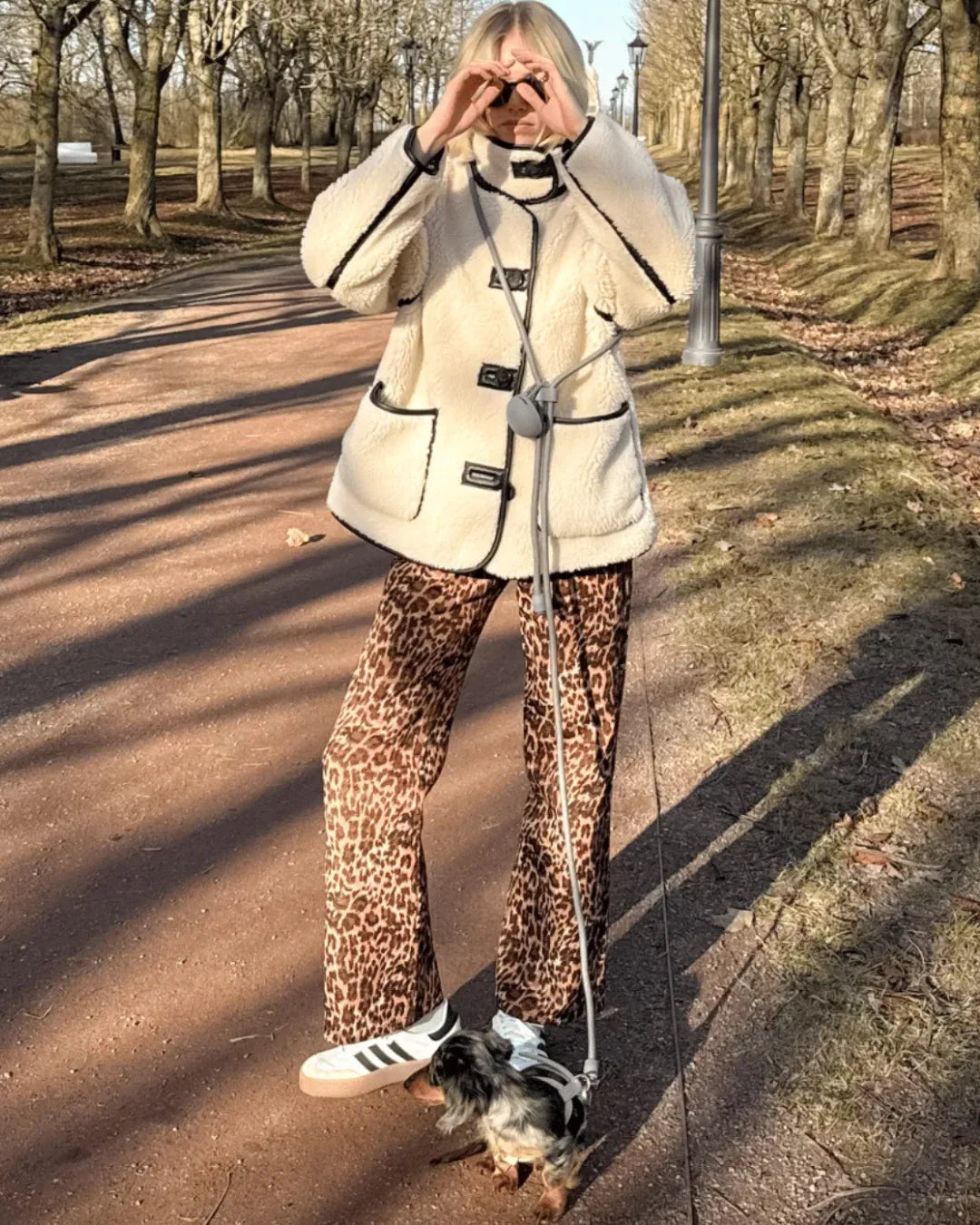 Person walking a dog on a grey handsfree lead in a park wearing a beige coat and leopard print trousers.