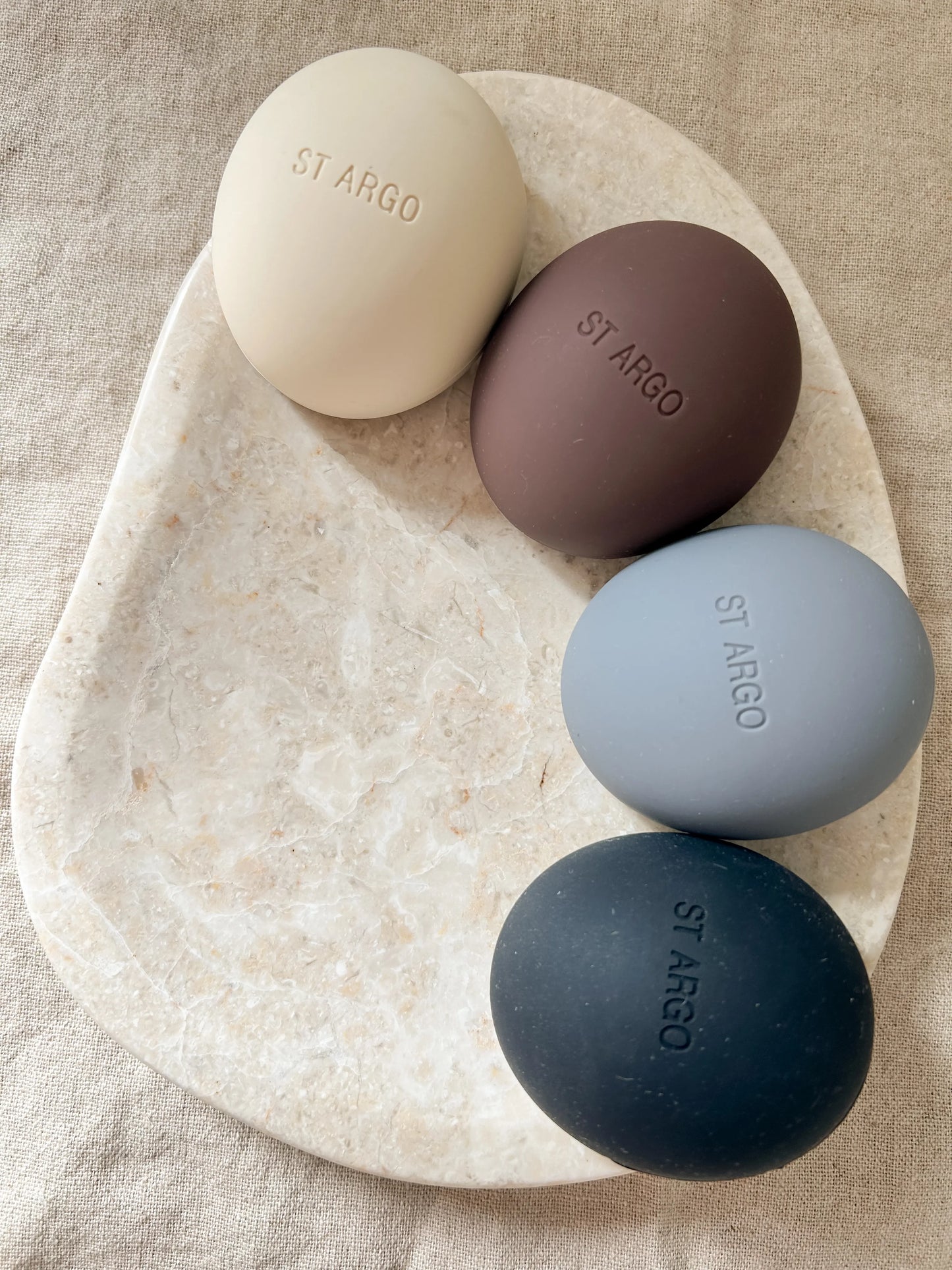 Four St Argo luxury dog treat pouches in beige, brown, grey and black on a neutral marble and cloth background