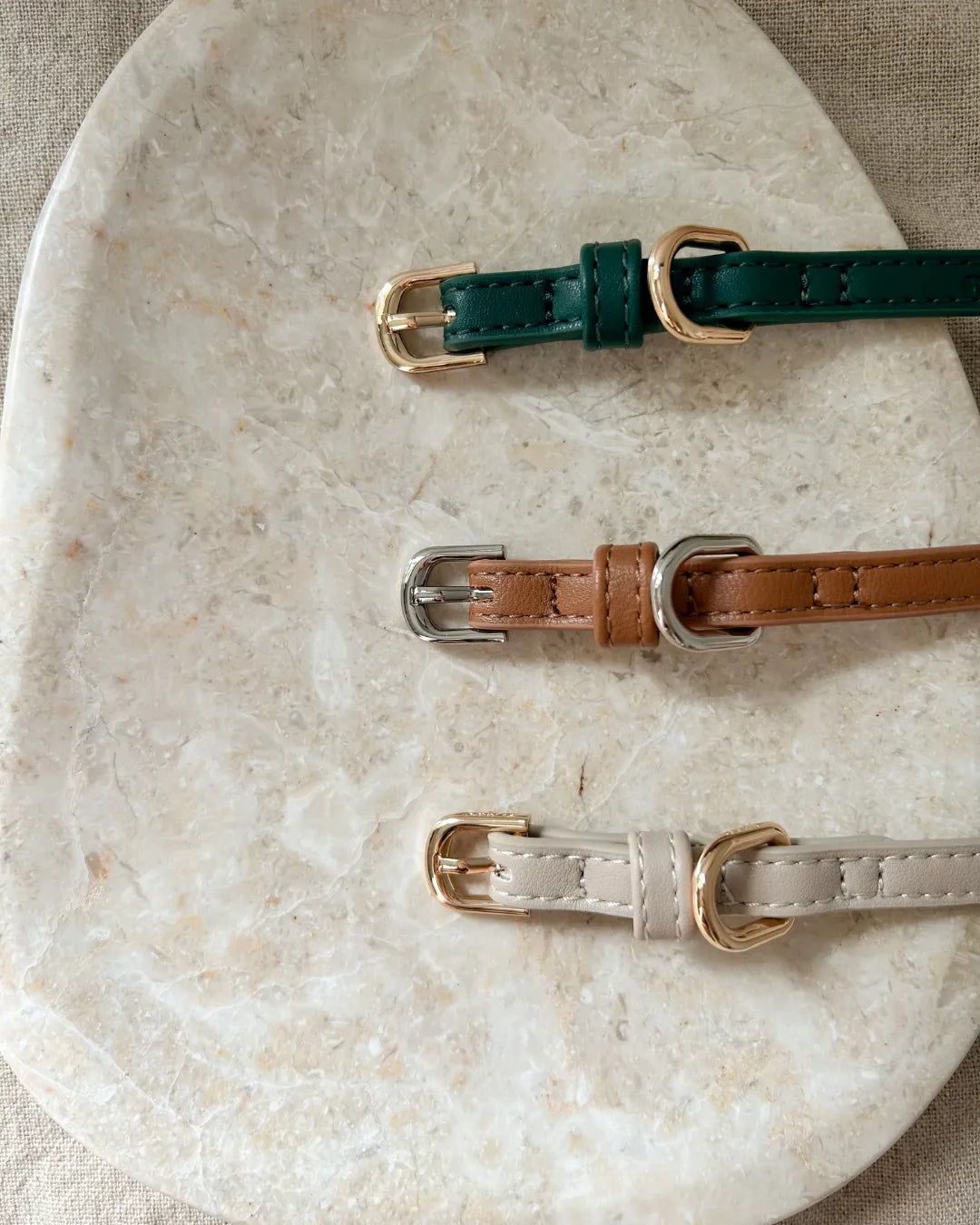 Three vegan leather dog collars in green, brown and beige on a marble surface.