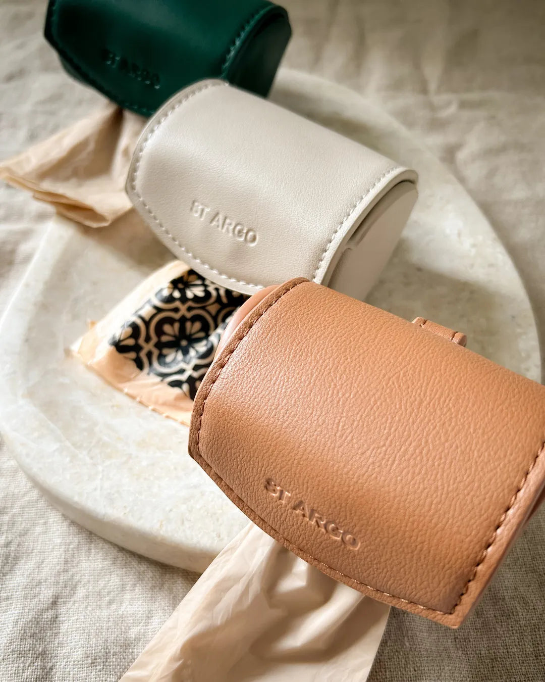 Close-up of three vegan leather dog waste bag holders with 'St. Argo' branding on a textured fabric background.