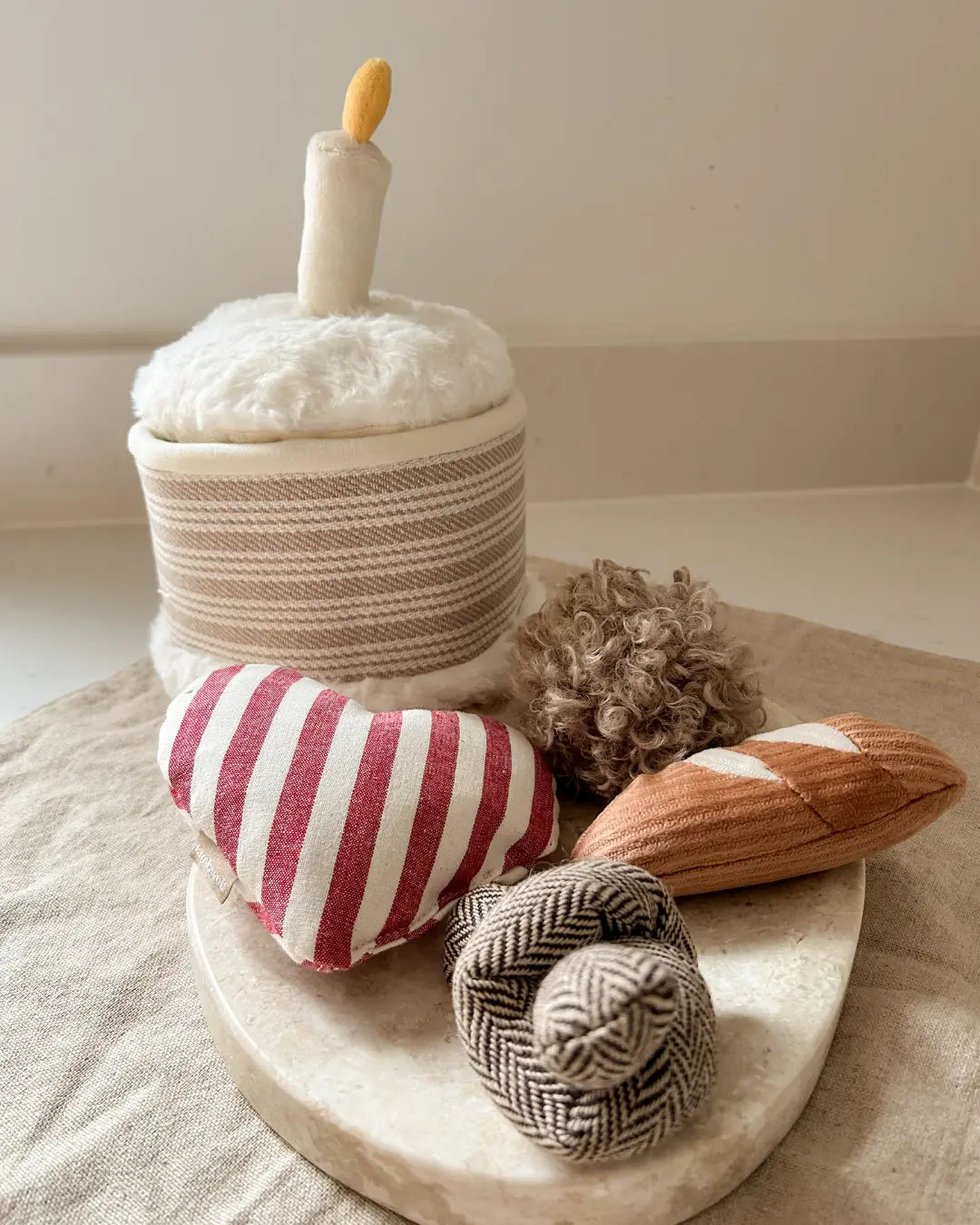 Plush birthday dog toys including a striped heart, textured ball, mini baguette, herringbone knot and fluffy cake on a neutral background