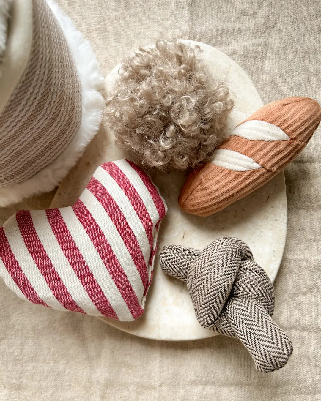 Plush birthday dog toys including a striped heart, a textured ball, a mini baguette and a herringbone knot on a marble surface