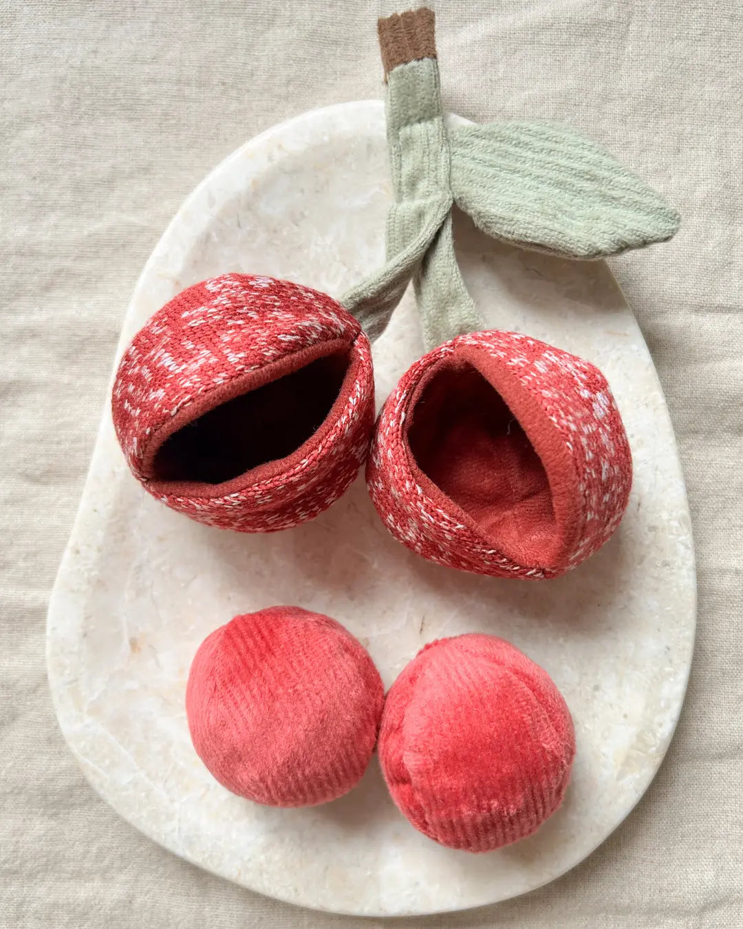 Red fabric cherry-shaped dog toys on a white plate with a leaf on a beige background