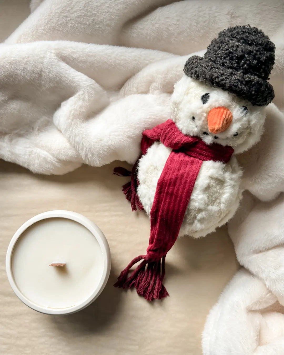 Plush snowman dog toy with a red scarf and black top hat next to a candle on a soft surface.