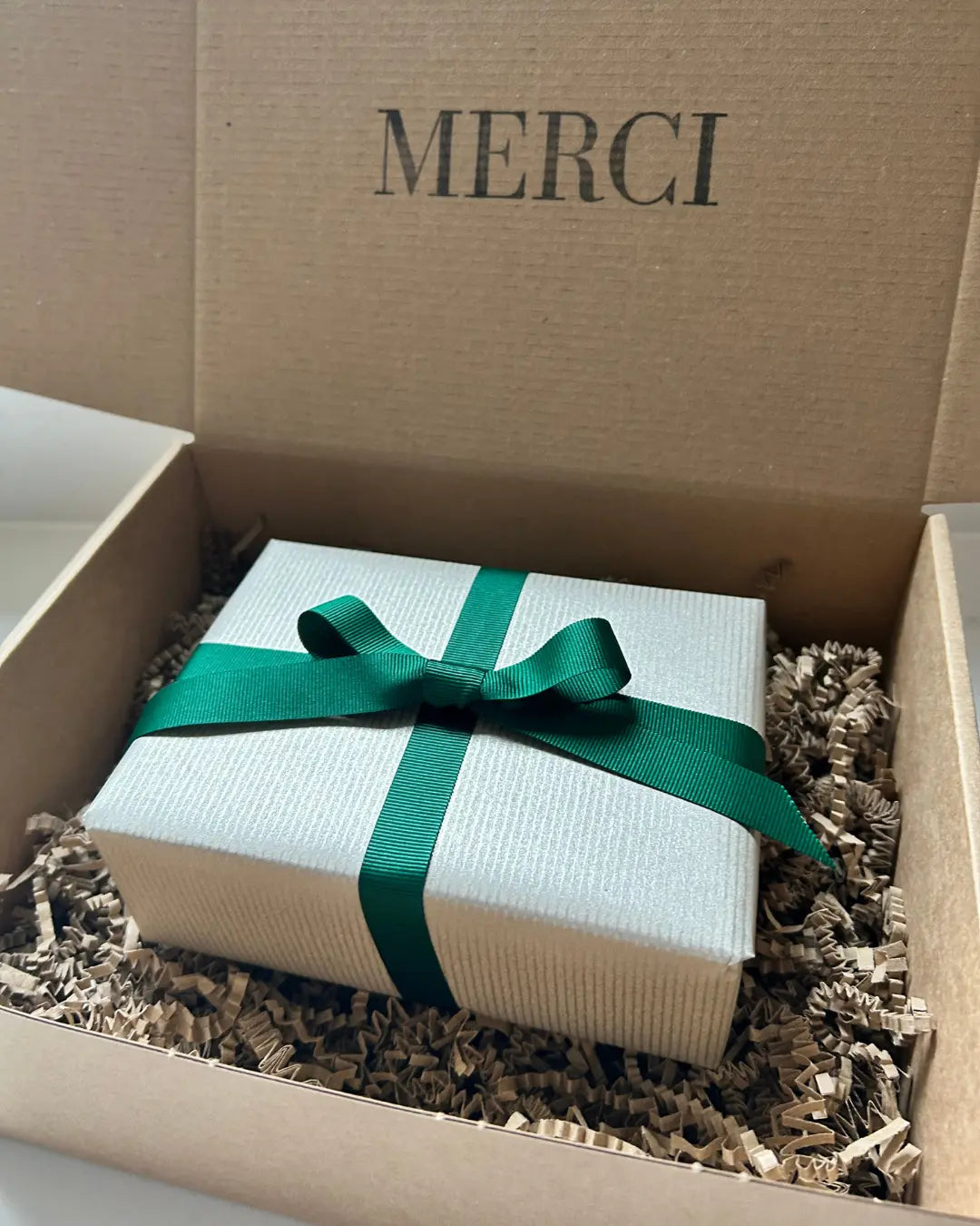 Gift box with a green ribbon inside a cardboard box labeled 'MERCI'.