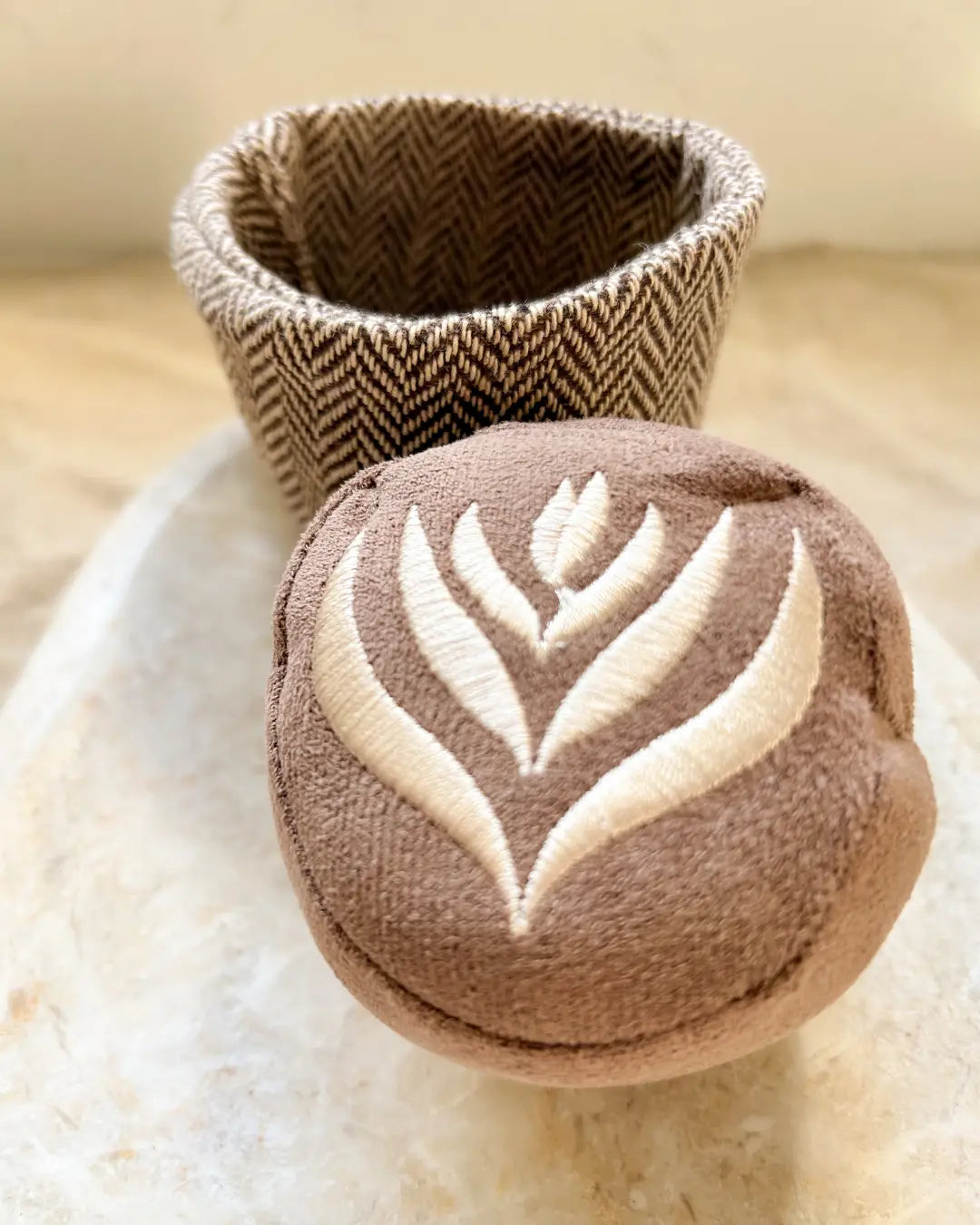 Plush dog toy in the shape of a coffee mug and a latte-patterned squeaky ball on a neutral background