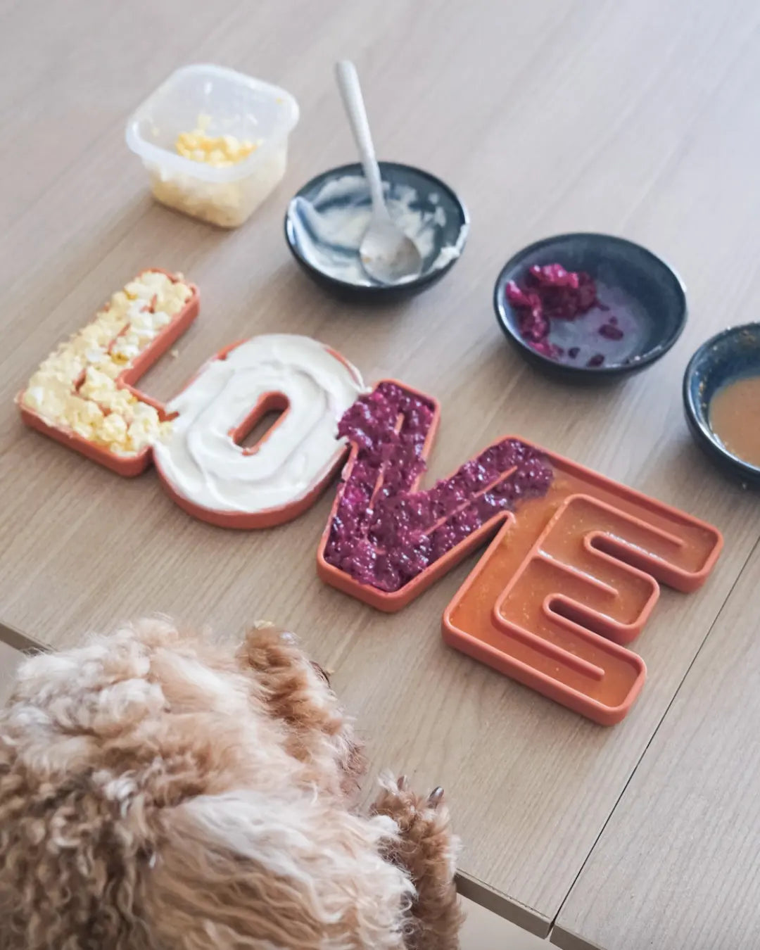 Dog lick mat shaped like letters spelling 'LOVE' on a wooden floor with a dog looking at them