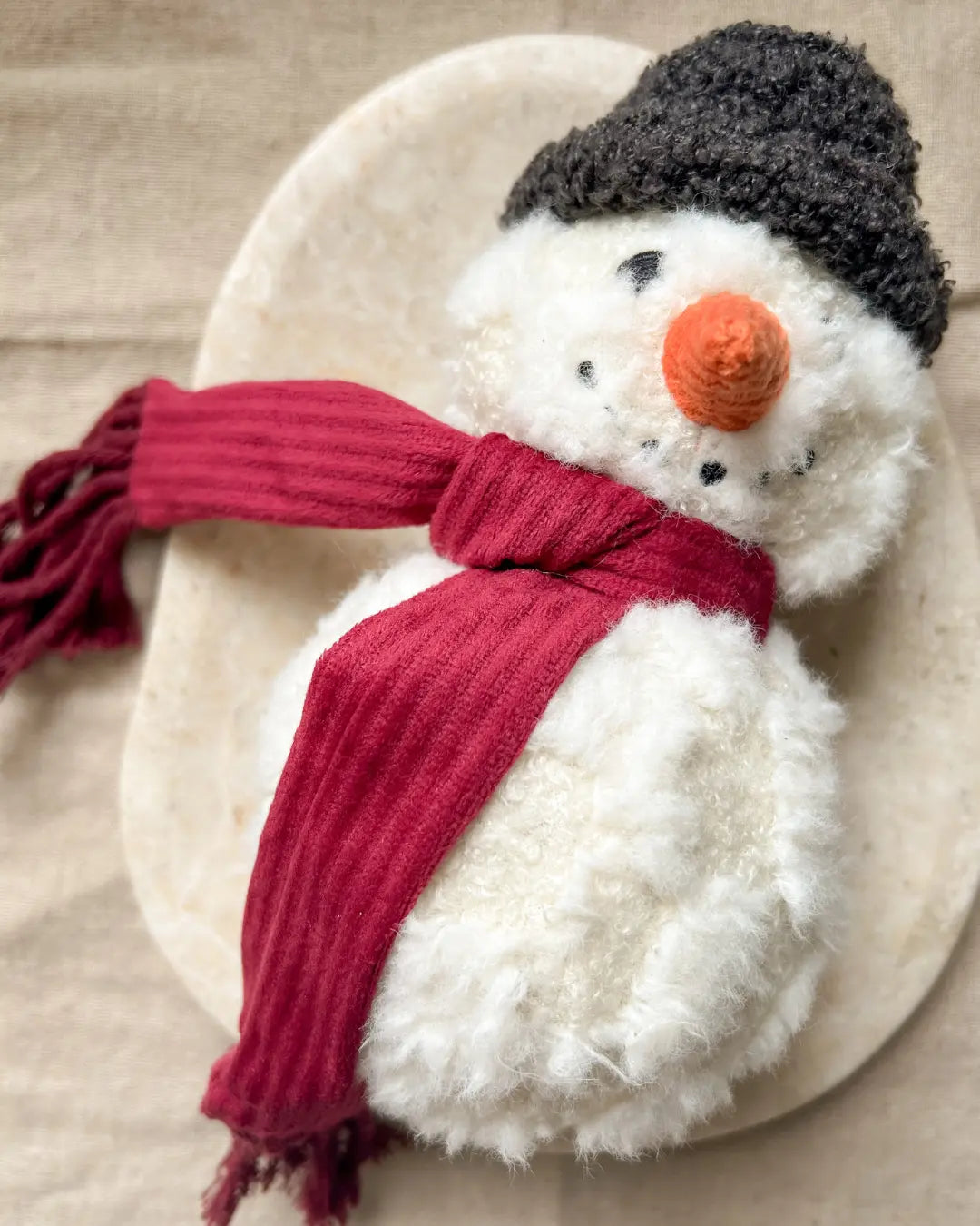 Plush snowman dog toy with a red scarf and black hat on a beige surface