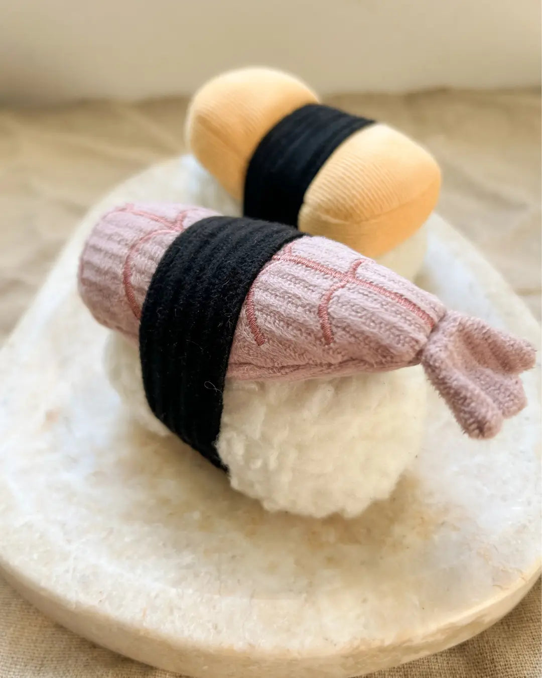 Two small sushi-shaped plush dog toys, one pink and one yellow, on a neutral background