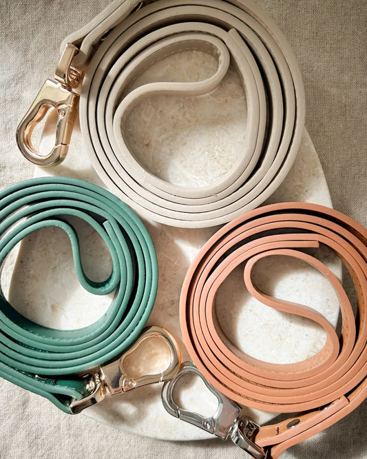 Three luxury vegan leather dog leads in beige, green and brown with metal clips on a textured surface.