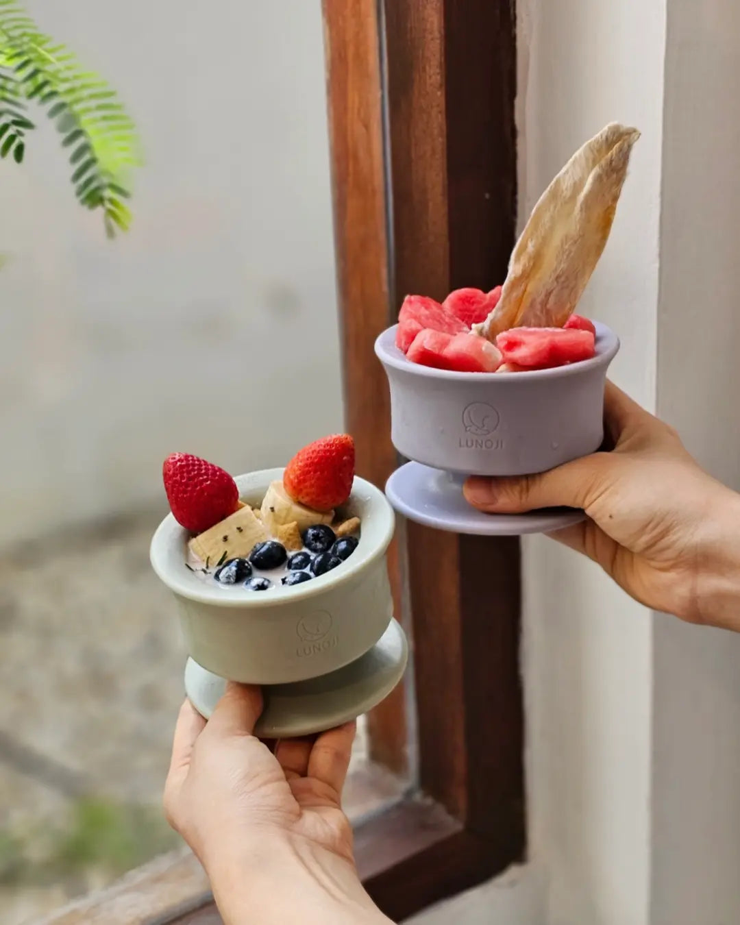 Two dog frozen enrichment bowls: one in sage green and the other in lilac, held in front of a window