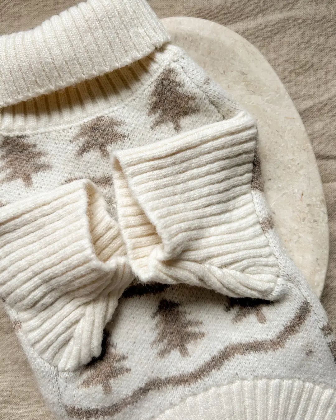 Close-up of a cream-colored knitted dog jumper with a Christmas tree pattern on a beige surface
